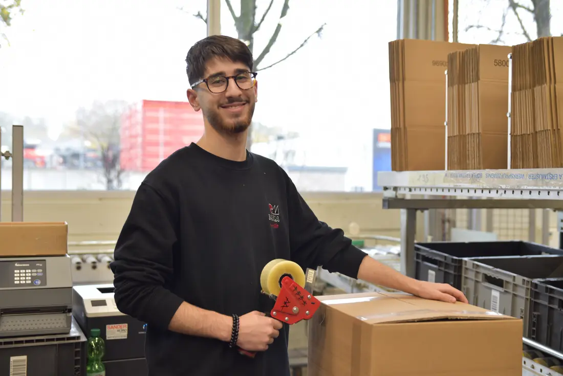 Ein Auszubildender steh am Arbeitsplatz im Versand und verpackt ein Karton. Er lächelt in die Kamera. 